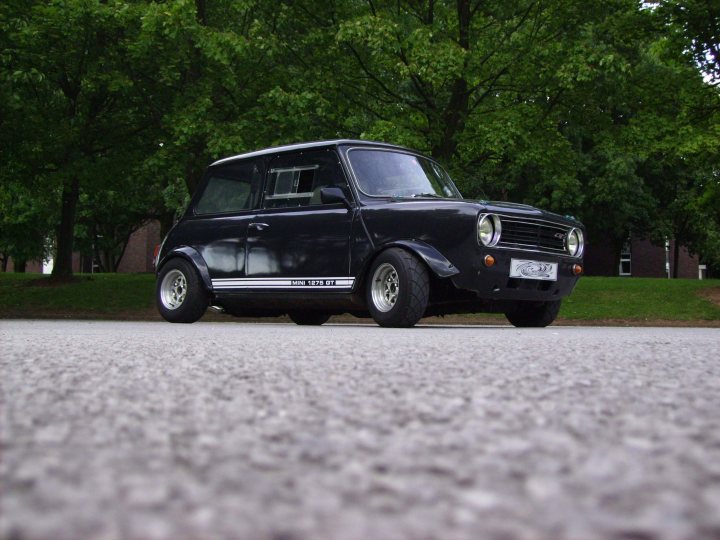 Get your car rated! - Page 29 - Readers' Cars - PistonHeads - The image depicts a small, dark-colored antique car parked on a gravel driveway. The car appears to be a two-door, possibly in the style of the 1970s or similar era. It has a round front shape, a traditional grille design, and classic rim wheel styles. The background shows a calm, tree-lined street and the greenery of a park or garden. The focus of the photo is the car, and the graininess of the shot, as well as the car's reflection on the gravel, suggest a shallow depth of field.