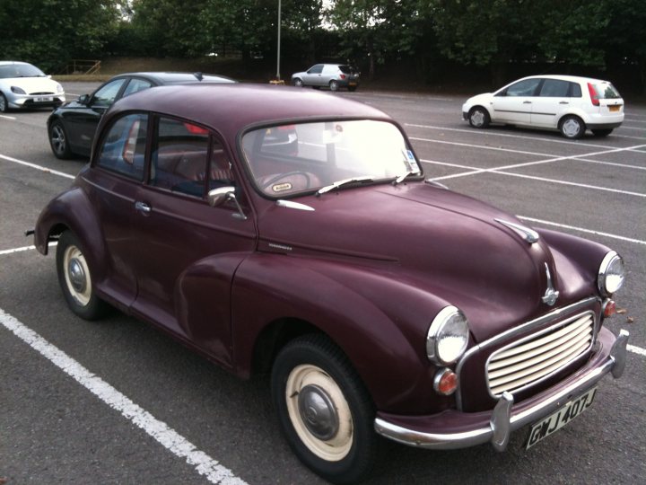 A car is parked in front of a fire hydrant - Pistonheads - The image features a maroon vintage car parked in a lot on a sunny day with clear skies. The car has visible damage to the hood, dashboard, and a passenger's side component. It is a 2-door vehicle with a compact size, featuring black wheels with a chrome hubcap. Additional details include a license plate located at the rear, and various outdoor elements like trees and other cars in the vicinity. The lighting suggests it is midday, with the brightest parts of the image being the car and the sky.