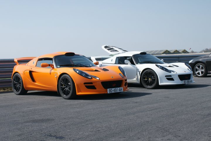 Chrome Orange Exige pictures please !! - Page 1 - Elise/Exige/Europa/340R - PistonHeads - The image is a color photograph depicting three sports cars parked on a paved surface at an angle to the camera. The foremost vehicle is a bright orange model with a sleek, low-profile design, featuring a prominent scoop on the hood and large air intakes near the sides. Behind it are two other sports cars, one white with a similar scoop and the other white with a more traditional silhouette without a hood scoop. All cars have their hoods opened, possibly for an event or show, and their black tires stand out against the pavement. This scene suggests an automotive event or car show, where high-performance cars are on display.