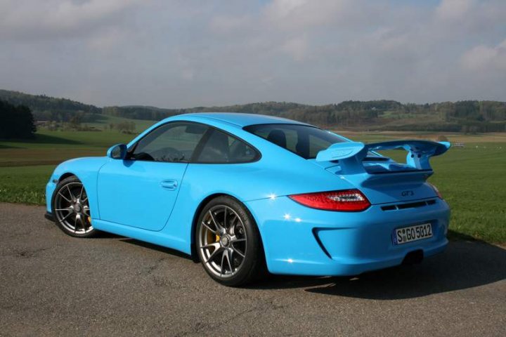 Pistonheads - The image depicts a vibrant, light blue Porsche GT3 in a grassy field. The car is positioned in a slight angle facing the camera, showcasing the iconic rear wing and the curved silhouette typical of Porsche sports cars. The vehicle's design and color make it a focal point against the natural backdrop.