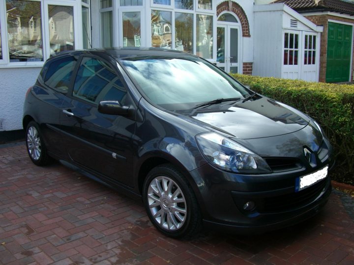 Midlanders Pistonheads - In the image, a dark-colored compact car is parked on a brick driveway. The car, a two-door hatchback, is positioned on the right side of the frame. The vehicle has a variety of aerodynamic features like a front air intake, black grills, and a spoiler for better aerodynamics. There is a clear reflection of the sky and clouds on its shiny, clean surface. The car sits in front of a house with a white exterior, and a small square top window which is likely a Velux window, providing natural light inside the dwelling. In the background, a well-maintained green hedge adds a touch of greenery to the white brickwork of the house.