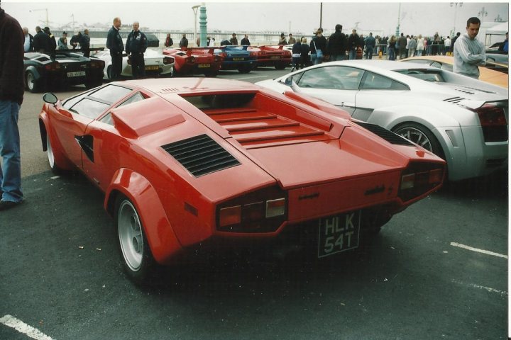 Countach  - Page 13 - Lamborghini Classics - PistonHeads - The image depicts a gathering of vintage cars in a lot. The most prominent car is a stainless steel red sports car with pop-up trunk, appearing to be the center of attention. Surrounding this car, there is an eclectic mix of other sports cars, predominantly in metallic shades like red and silver. People can be seen admiring the vehicles, suggesting this could be a car show or exhibition. The setting appears to be an outdoor car lot with ample space for the cars and spectators. The overall atmosphere is casual and leisurely, indicative of a public display or event.