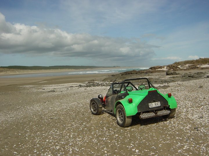 Beach Pistonheads Mile - The image depicts a vintage-style, convertible vehicle being driven on what looks like a pebble beach. The car is painted in a color scheme that at first glance suggests it may be green, but upon closer inspection, reveals the familiar pattern of white seashells. The beach itself appears to be calm with whitecaps visible on the ocean in the background. The sky is mostly cloudy, providing a dramatic backdrop to the scene. The overall atmosphere suggests a leisurely or adventurous drive along the coastline.