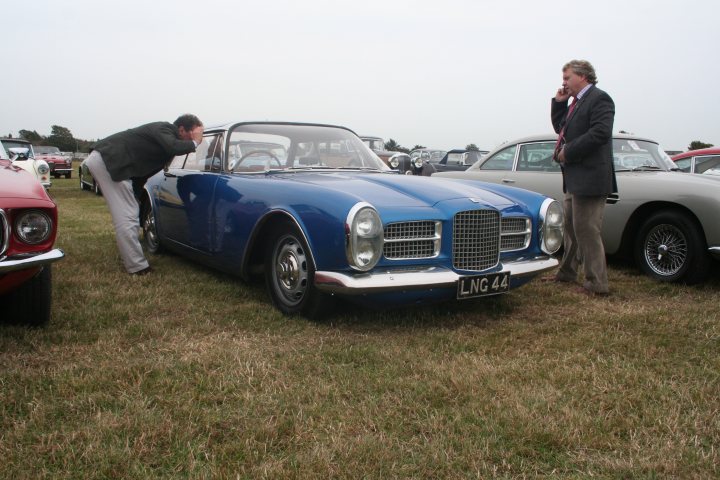 Vega Facel Pistonheads - This image captures a tranquil scene in a grassy field where several cars are parked. The focus of the image is a man dressed in a suit and tie, standing next to a striking blue vintage Bentley car. His hand is raised to his ear, suggesting he might be on the phone. The car's vintage design stands out among the other cars parked nearby. The setting is serene, with the cars parked in an orderly fashion on the grassy field, and there are trees and other cars in the background, adding depth to the scene.