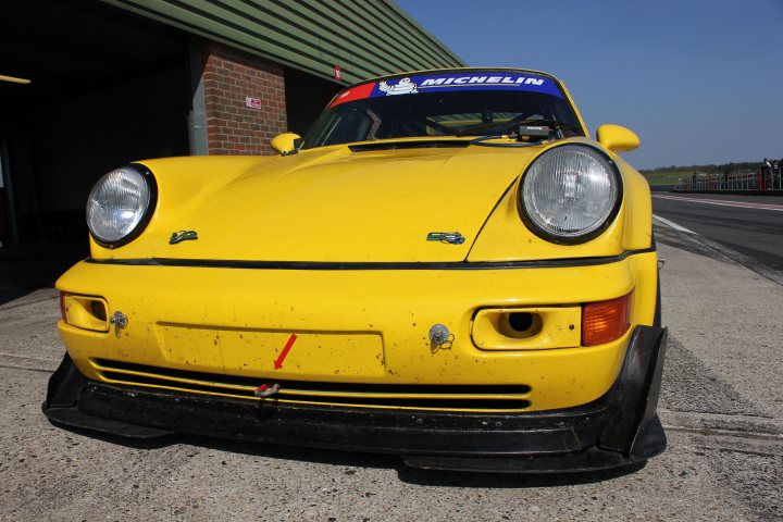 The annual what are you racing in 2014 thread.  - Page 1 - UK Club Motorsport - PistonHeads - The image features a bright yellow sports car parked outside a garage with a green metal roof. The vehicle has distinctive blue and white markings, which include the word "MICHELIN" and a cartoon character design. The car is positioned to show a close-up view of its front, complete with angled headlights and a pointed license plate. The license plate has a red horizontal line, which stands out against the black and yellow license plate holder. The setting is a clear, sunny day, indicating that the photo was taken during the daytime.