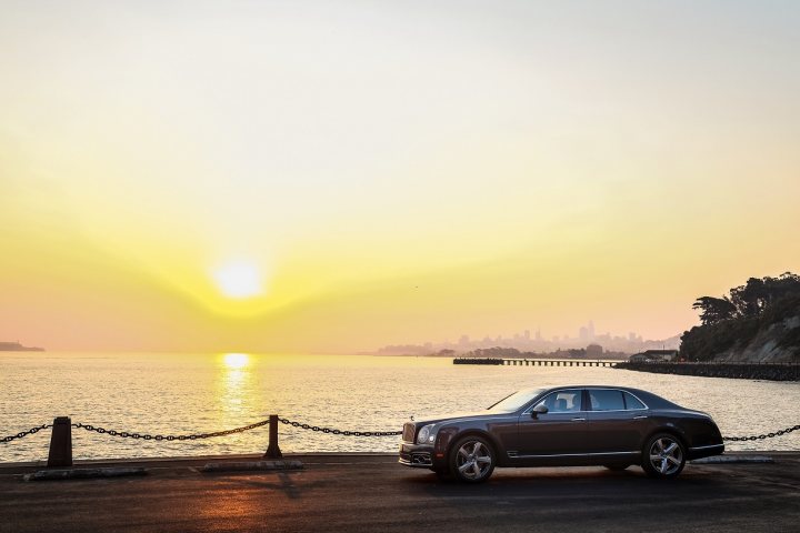 Regular Mulsanne or Speed - Honest advice please? - Page 5 - Rolls Royce & Bentley - PistonHeads - The image captures a serene scene at sunset. A black luxury car, glossy and sleek, is parked on the side of a road adjacent to a body of water. The car's sleek profile contrasts with the rough texture of the concrete road. The road is bordered by a chain, adding a sense of security or restricted access to the scene. Beyond the road, a large body of water stretches out, its surface reflecting the warm hues of the setting sun. In the distance, the faint outlines of a city skyline indicate the presence of civilization on the other side of the water. The sky is painted with soft, golden hues, creating a warm backdrop to the scene. The overall atmosphere of the image is one of tranquility and luxury.