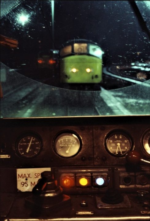 Loco sheds and other railway buildings.... - Page 18 - Boats, Planes & Trains - PistonHeads UK - The image displays a split-screen of two views related to train travel. In the top half, there is an interior view from inside a passenger car on a train, showing the side window with reflections, including a lit street lamp and another train car in reflection. It appears to be nighttime or early evening due to the darkness outside the vehicle.

In the bottom half, we see an exterior perspective featuring the front of a train at a station. The train has a distinctive green color and is equipped with classic railway gauges on the left side. This suggests that this is likely an older model or designed to have an antique appearance. There are indicators for speed and track information, including directional signals and a warning sign.

The photograph has been edited together from two separate images, as evidenced by the visible split between the top and bottom frames. The style of photography is realistic with attention to detail in the reflections and the mechanical components of the train, emphasizing the setting within a railway context. There are no texts visible in this image.