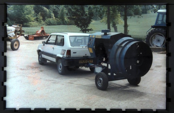 Fiat Panda 4x4 Mk 1 Fire.  A  Classic Car - Page 1 - Classic Cars and Yesterday's Heroes - PistonHeads - The image captures a scene of a white hatchback car parked on a concrete surface, partially hecked by a large blue metal object with circular parts. To the left of the car, a plough tractor is visible in the background, suggesting an agricultural setting. On the right side of the frame, there's another object, possibly a piece of machinery, partially blocking the view of the tractor. There's a person in the background, standing close to the tractor. The ground around the car and the parked vehicle is sparse, with some grass and trees visible nearby. The overall color palette of the image is earthy, with the blue, white, and green standing out against the more neutral tones of the concrete and ploughed earth.