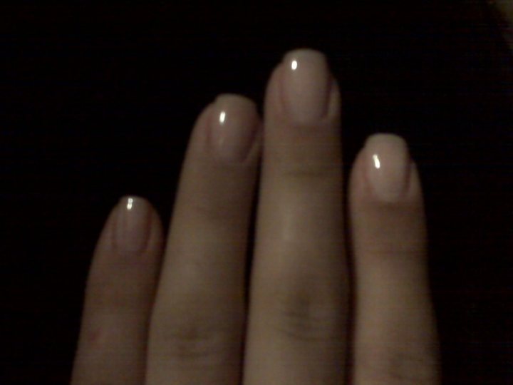 The image shows a close-up view of two hands, with a focus on the nails. The nails are well-groomed, with a light pink glossy polish. The hands are holding a part of a garment, and the background is dark, which suggests that the image might have been taken in low light conditions. The fingernails are shiny and clearly manicured, reflecting a certain level of care for personal appearance. The image composition emphasizes the hands and nails, creating a clear focal point for the viewer.