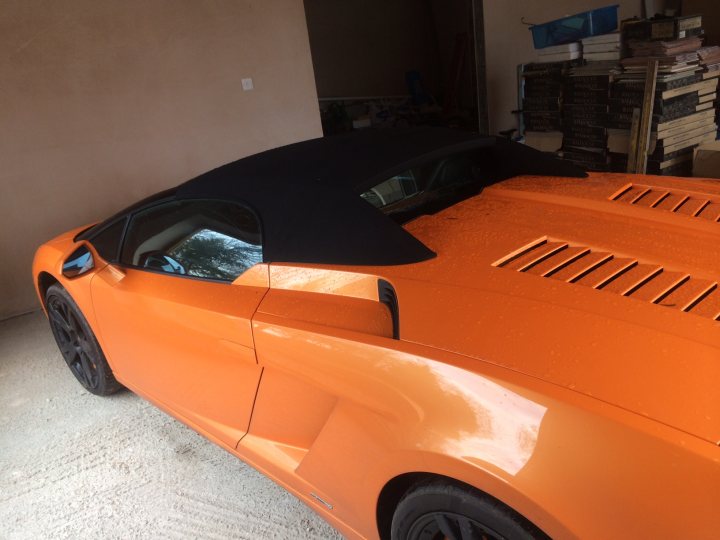 More pics of my LP560 - Page 1 - Gallardo/Huracan - PistonHeads - The image shows a section of a bright orange sports car parked inside a garage with its convertible top down, revealing a black interior underneath. The car is a convertible, as indicated by the exposed interior. In the background, there is a collection of books stacked on the floor. The car's design is sleek, with a high gloss finish that reflects the ambient light. The image is taken close to the car, capturing the bottom half of the vehicle and a portion of the interior.