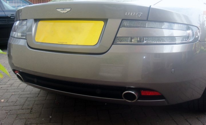 Clear Rear Lights Install - Page 1 - Aston Martin - PistonHeads - The image shows the rear of a silver sedan parked on a paved surface. The vehicle is viewed from a slight angle, with another indistinct vehicle visible on the left side of the frame. A prominent feature is the yellow license plate frame attached to the license plate, which contrasts with the car's color. The vehicle appears modern, with a clean design and sleek lines.