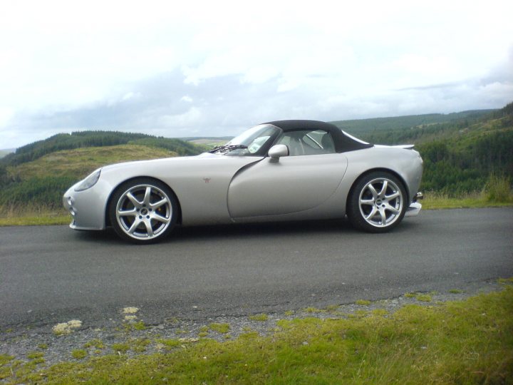 Some pictures of my Tam - Page 1 - Tamora, T350 & Sagaris - PistonHeads - This image features a silver, convertible sports car parked on an asphalt road with grassy areas at the sides of the road. The vehicle is sleek and shiny, suggesting it's well-maintained. The car is equipped with a large spoiler at the rear and five-spoke alloy wheels. In the background, there's a scenic landscape with hills covered in a mixture of grass and trees, creating a serene and picturesque environment. The weather appears overcast with a cloudy sky.