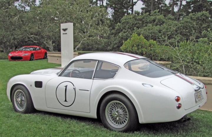 Pics from Aston event, Monterey - Page 1 - Aston Martin - PistonHeads - This image features a vintage white car parked on a grassy field. The car is adorned with a "1" on its side, suggesting it might be from a production or racing series where the number is significant. In the background, there is a red sports car and a building that says "Cobra" on it. The overall scene appears to be a classic car show or a museum dedicated to vintage automobiles.