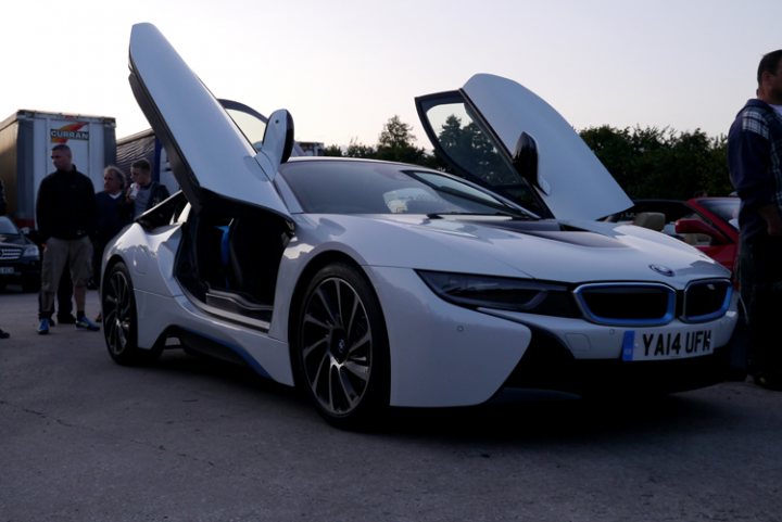 The GT Cafe - the ace cafe of the north - Aug meet - Page 1 - North West - PistonHeads - This image captures a scene at a parking lot or garage. The main focus is on a white BMW i8 sports car, which is parked near the center of the image and occupies a significant portion of it. This sports car is known for its distinctive and somewhat controversial carbon fiber door panels that increase visibility when the car is parked with its doors open, as seen in the image. In the background, there are other cars and people, adding to the ambiance of a busy location. The setting suggests the car might be on public display or at an event, as indicated by the attentive spectators scattered throughout the scene.