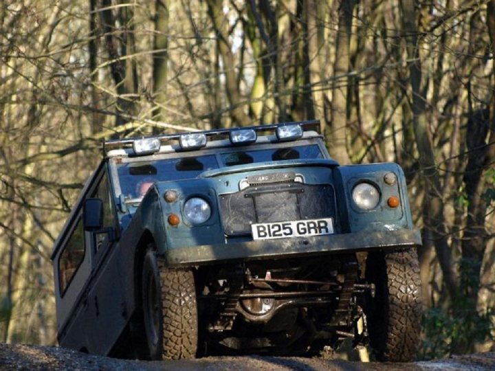 Hampshire Lanes Road Pistonheads - The image shows a scene featuring a robust off-road vehicle, potentially a Land Rover, positioned on a muddy path surrounded by dense foliage indicative of a forested area. The vehicle has a visible license plate that reads "B125 GAR." The front of the vehicle is inclined, a characteristic of off-roader models built to traverse uneven terrain. The tire treads appear well-worn, suggesting frequent use in such conditions. The overall setting and the vehicle's attributes imply it's capable of navigating in diverse natural environments, designed for outdoor adventure.