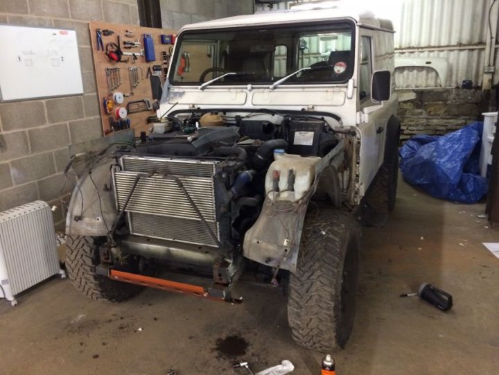 Land Rover 90 TD5 Tribute Build & Video Series - Page 1 - Readers' Cars - PistonHeads - The image shows a white off-road vehicle parked in a workshop with a concrete floor. The vehicle appears to be a utility or police truck, given its rugged design and the arrangement of its parts. The engine is out of the vehicle, indicating some maintenance or repairs are being carried out. On the back of the vehicle, there is a large, vertical metal grill that likely serves as a bumper or defensive mechanism. The vehicle is predominantly white, with no visible texts or logos that could indicate its brand or intended use.