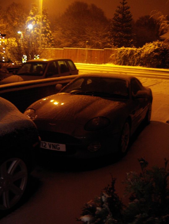 The *AM in the snow* thread :) - Page 2 - Aston Martin - PistonHeads - The image is a nighttime photograph of a snowy scene featuring a car parked on the side of a street illuminated by street lights. The car, which appears to be a Jaguar, is positioned in the foreground of the image, facing the camera with snow visible on the ground and the car itself. 

The street is lined with a fence made of wooden slats, and the sky visible in the background suggests it is either early morning or late evening. The lighting from the vehicles on the left and the street lights contribute to the wintery ambiance of the scene. 

There are no visible texts or distinctive marks that provide additional information about the location, event, or context of the image. The composition concentrates on the car and the immediate vicinity, offering a quiet, wintry atmosphere.