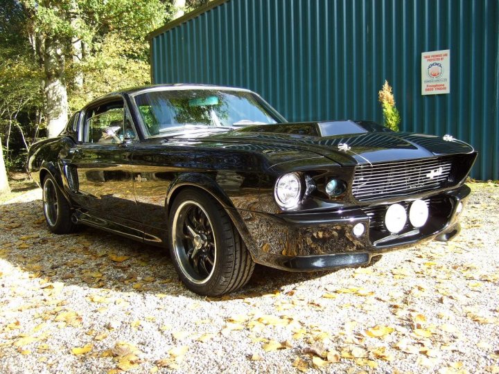 Coolest background car ever.....(my humble opinion) - Page 1 - General Gassing - PistonHeads - The image shows a black, modified, muscle car with a scooped hood, parked on a gravel surface close to a building. The car has white-wall aftermarket rims, and a dual Liberty headlight is prominently visible. In the background, there are trees and a blue metal wall. A sign indicating "No Heavy Goods Vehicle" is visible on the wall behind the car. The overall style of the image suggests a photograph taken during the day with natural light highlighting the car.