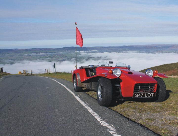 Lotus 7 - S4 - Page 7 - Caterham - PistonHeads - The image captures an iconic scene of a red and black Lotus Seven on a winding mountain road. The car, known for its agility and lightweight design, is parked on the side of the road with a mountain's peak visible in the background, suggesting a high-altitude location. Above the car, a red flag is fluttering on a pole, while another flag and a classic wooden gate are also visible on the road. The overall setting is serene and open, offering a glimpse into the beauty of the landscape.