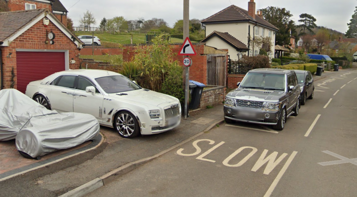 misbadging cars - Page 187 - General Gassing - PistonHeads UK - The image is a photograph showing a residential street scene. There are parked cars on the side of the road, including a white sports car and a few other vehicles. A parking sign is visible near the curb with the word "CLOSED" in bold letters. On the right side of the photo, there's a blue car driving away from the camera. The street appears to be in an urban or suburban area, as evidenced by the presence of houses and other typical street elements.