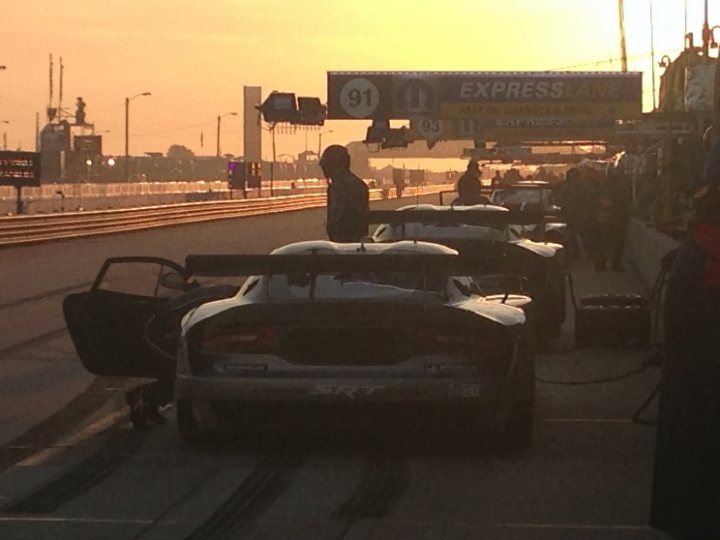 New SRT Advert - Page 1 - Vipers - PistonHeads - The image portrays an active scene on a race track just as the sun is setting. Numerous sports cars, possibly Corvettes, are lined up in the pits, their sleek designs and dark colors stark against the golden hue of the sky. A person, likely a pit crew member, is standing by a car, perhaps attending to it before the race begins. There are a few signs visible in the photo, but the specific details are not clear. In the background, the setting sun paints a stunning contrast against the silhouetted cars and towering grandstand in the distance.