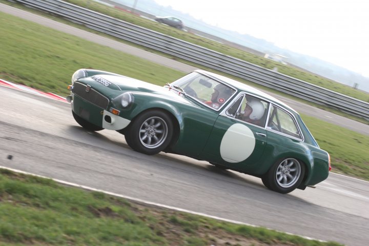 MGB or MGC? - Page 1 - MG - PistonHeads - The image features a distinctive green racing car on a racetrack. The car exhibits a vintage design reminiscent of classic European sports cars, with a white oval patch on its body, possibly a sponsor's logo or a racing number. The vehicle is in motion, as indicated by its position in the frame, with its front tires leading the way. It is daytime, and the racetrack appears to be outdoors with a grassy area along one side. The background shows some hills or a gentle slope, adding a natural element to the racing context.