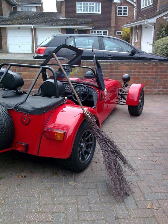 Parking Close Pistonheads - The image depicts a vibrant red car with a unique feature: a large broom attached to the back. Lying on the car is a black gas tank, possibly used during the day. The car is parked in a brick parking lot adjacent to a residential area with houses visible in the background. The weather appears to be overcast, suggesting it might be a cool or cloudy day. The overall scene has a playful and whimsical vibe.