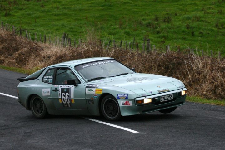 Race Rally Pistonheads Sprint - The image features a racing car captured in motion on a paved road. The car is a sports coupe with a prominent rear spoiler and a decal on the side door displaying the number '66', which is often associated with racing. The car's body has a duco paint finish, and it's outfitted with racing stickers and decals, reinforcing its competitive nature. The background of the image shows the side of a grassy hill under a clear sky, indicating an open-air countryside setting. The car's dynamic pose and the blurred background suggest it's moving at considerable speed, adding a sense of motion and thrill to the scene.