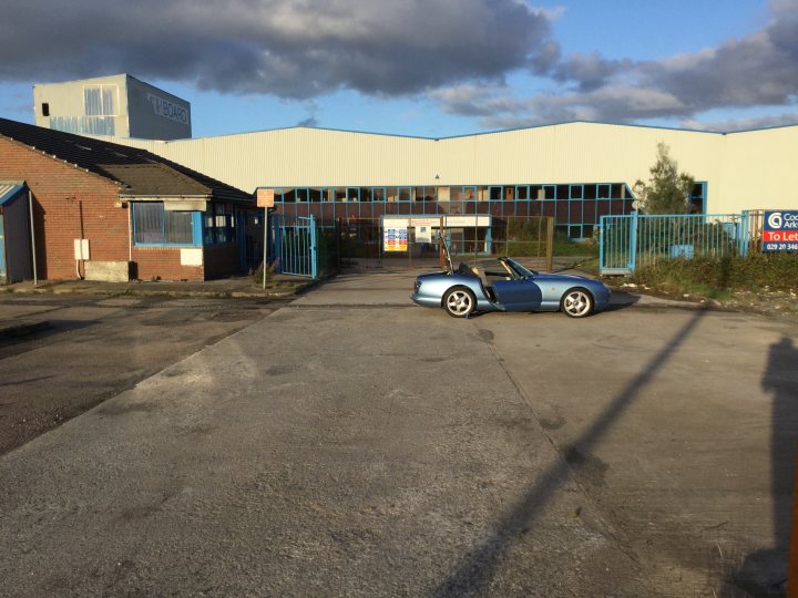 New TVR still under wraps! - Page 27 - General TVR Stuff & Gossip - PistonHeads - The image features a road with a blue car parked on the side. There is a brick building with a corrugated metal roof in the background, along with what appears to be an industrial or commercial structure. The ground is flat with minimal details, and the sky is overcast with a grayish tone. There is no text visible in the image.