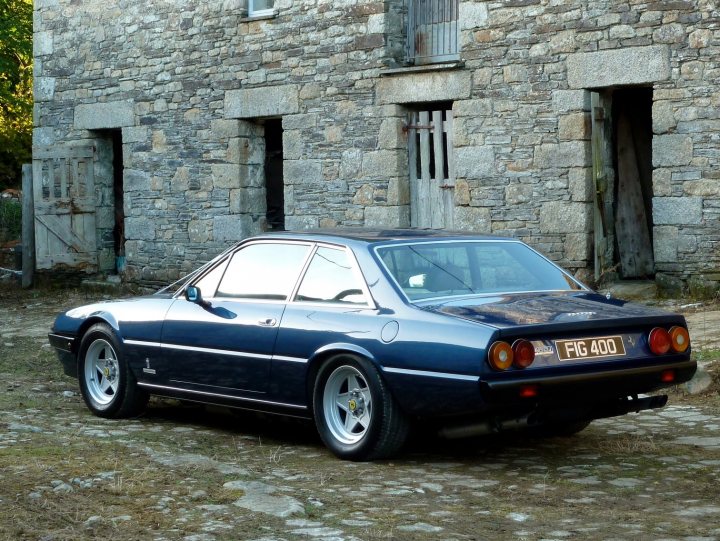 A mate's new toy - Page 1 - Ferrari V12 - PistonHeads - The image portrays a classic, dark-colored coupe parked in front of a stone building. The car appears to be an older model with distinctive round headlights and a distinctive rear design, indicative of models from the 70s or 80s, which seems to be sitting in a gravel-covered driveway. The stone building has a rustic look with wooden doors and sloping rooflines, adding to the overall vintage charm of the scene. On the driver's side of the car, there's a visible license plate that reads "FG400". The contrast between the sleek gray vehicle and the rugged stone structure creates a visually appealing scene that speaks to classic automotive aesthetics.