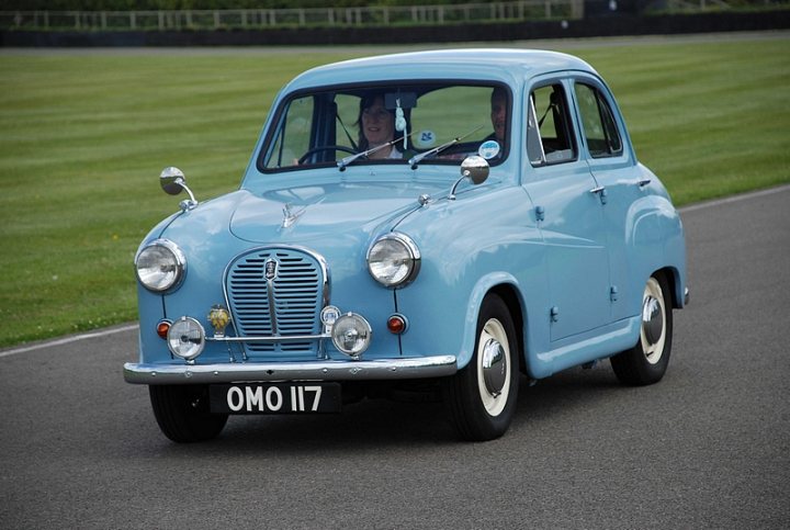 Goodwood Breakfast Club, Pre 1966 classic sunday - Page 1 - Goodwood Events - PistonHeads - The image shows a vibrant, light blue vintage car parked on a paved road. The car appears to be in motion, as suggested by its license plate reading "OMO 117." It has a split windshield typical of vintage cars, and the grille and headlights mirror the classic design of a bygone era. The vehicle is parked on a road with well-trimmed grass and other vehicles in the background, suggesting a public or communal parking area. The image has a clear, focused composition that emphasizes the car as the main subject.