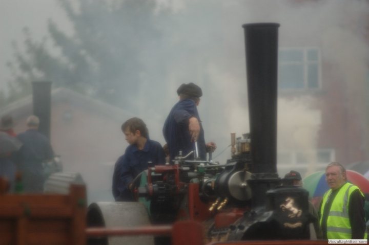 Classic tractors - Page 3 - Classic Cars and Yesterday's Heroes - PistonHeads - The image portrays an outdoor scene where a vintage steam tractor is being operated. Two men, dressed in blue jackets, are at work on the tractor, with one person being at the engine level while the other is standing above it, possibly adjusting or operating the procedures necessary to produce steam. One of the individuals is also holding a tool, indicating maintenance or operational activities.