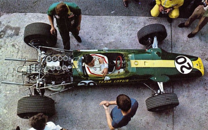 Pistonheads - In the image, the main focus is a vintage Lotus Formula 1 racing car, resting on a concrete surface. The car is painted in a striking mix of green and yellow, with the iconic black and white number 20 prominently displayed on the side. There are four individuals present in the image. A man, presumably the driver, wearing a white suit is seated inside the cockpit of the Lotus, appearing to be gazing towards the rear. Three other men, dressed in green and white, are in the vicinity of the car, possibly part of the crew or team members, as one of them is holding a camera and another seems to be adjusting a component on the car. The image conveys a sense of anticipation and preparation, typical of a motorsport event.