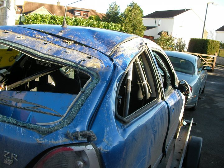 Where is everyone?  - Page 1 - MG - PistonHeads - The image displays a blue car in a parking area, with part of its rear window missing, exposing the interior. The car has a noticeable crack across the back window. The car also shows speckles and debris on the top of the vehicle, suggesting it may have been in use or had an accident. In the background, there is another car parked, and elements of a residential street visible, including a hedge and a house. The photo appears to be taken during the day under natural light.