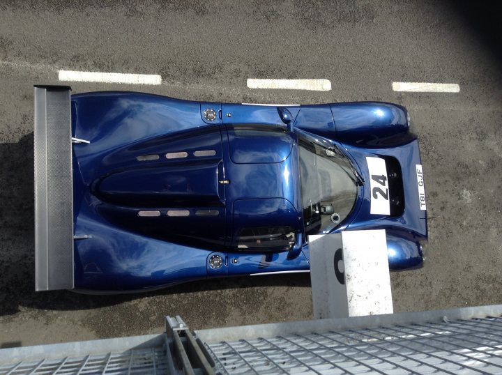 GRRC Spring Sprint at Goodwood - Saturday 10 May - Page 1 - Goodwood Events - PistonHeads - This is a photograph of a single-seat sports car from an elevated angle. The vehicle is predominantly blue with aerodynamic bodywork and a large rear wing to generate downforce. The car is equipped with a covered number 24 that appears to indicate its racing number, and a round badge that might be a manufacturer's logo on the front side panel. There is a visible driver seat with a racing harness, hinting at its purpose as a competition or racing vehicle. The tarmac is divided by white lines, suggesting a predetermined area, possibly a track, where the car is being stored or used.