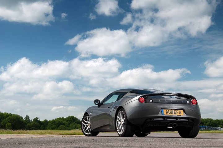 Evora at speed  - Page 1 - Evora - PistonHeads - The image captures a scene of a black Audi R8 sports car parked on the side of a road. The car is facing towards the right side of the photo, and it has a license plate that reads "AUI E7A". The weather in the photo is cloudy, suggesting an overcast day. The road on which the car is parked seems to be in a rural or semi-rural area, with greenery visible in the background. The overall composition of the image suggests a peaceful setting, creating a contrast between the stillness of nature and the dynamic potential of the sports car.