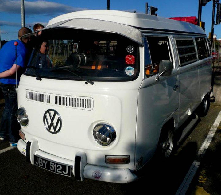 MetroCentre pics - Page 1 - North East - PistonHeads - The image shows a vintage Volkswagen bus parked on what appears to be the side of a road. The bus is predominantly white with some black detailing and has a distinctive round emblem on the front. It has a large rear window and side windows, along with side mirrors. There are stickers and what looks like a license plate visible on the front. In the background, there are two people, and to the right, an umbrella is partially visible, suggesting a recent rain or the impending possibility of rain. The lighting suggests it is daytime.