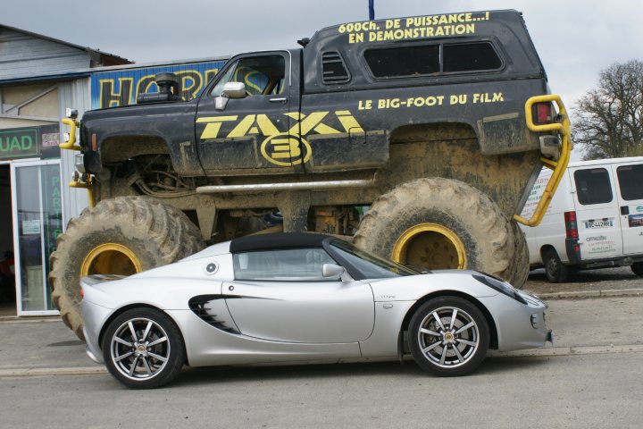 Pistonheads Size Doesnt Matter - The image shows a large truck with a massive front tire and tracks, towering over a smaller silver car parked in front of it. On the truck, there is a sign advertising "Taxis," and other text is present, including "MONTAGNE," "TAXI," "BIGFOOT," and "DU FILM." A van is partially visible in the background on the right side of the image. The setting appears to be outdoors during the daytime. There are men standing near the vehicles, but their bodies are partially hidden from view, making attire and full identities indeterminable.