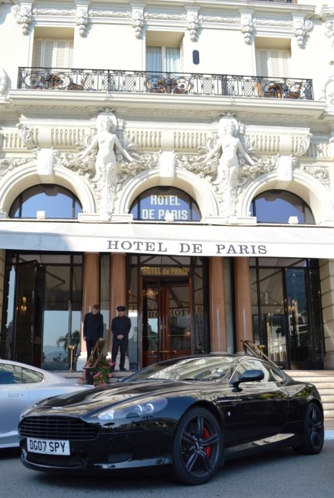 Show us your DB9  - Page 3 - Aston Martin - PistonHeads - The image features a sleek, black Aston Martin car parked in front of a grand building that is the Hotel de Paris in Paris. The hotel's facade is white and intricately designed with columns and carvings, suggesting a luxurious and historical establishment. There are two individuals standing on the steps in front of the hotel; one is wearing a black suit and the other is wearing a uniform, possibly indicating that they are staff or security for the hotel. The sky is overcast, and the overall feel of the image is one of elegance and sophistication.