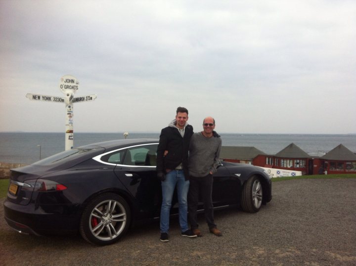 Land's End to John O'Groats electric car records - Page 1 - EV and Alternative Fuels - PistonHeads - This image shows two men standing in front of a black sports car, which is parked on a gravel surface. Behind the car is a black and white sign featuring encounters along a coastline. The sign indicates points of interest such as John's Ledge and O'Groat's Ferry. The weather appears overcast, and the men are dressed in casual attire. The scene suggests a scenic location near the coast.