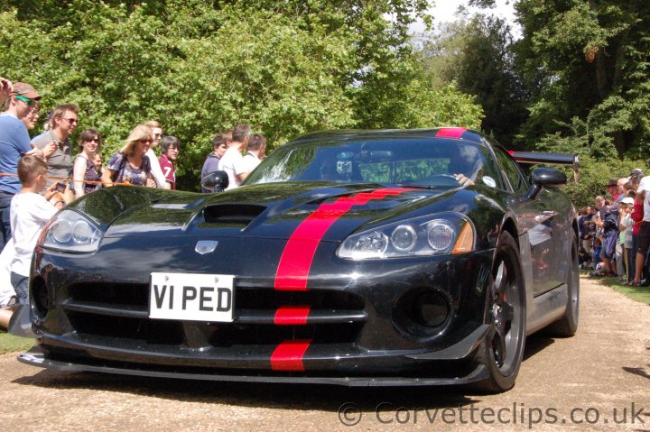 wilton house pics - Page 1 - Vipers - PistonHeads - The image captures a dynamic scene at a car show. Dominating the center is a striking black and red Viper sports car, its sleek design and vibrant colors drawing the eye. The car is stationed on a dirt road, adding a rustic charm to the scene. 

Surrounding the car is a crowd of people, their expressions suggesting excitement and admiration. Some are standing closer to the car, perhaps appreciating its design or contemplating what it might be like to drive it. Others are a bit further away but still close enough to capture the details of the car and the expressions on their faces. 

The crowd and the car together create a sense of anticipation, as if something remarkable or entertaining is about to take place. The precise positioning of the objects suggests an organized event, with the car on display surrounded by an engaged audience.