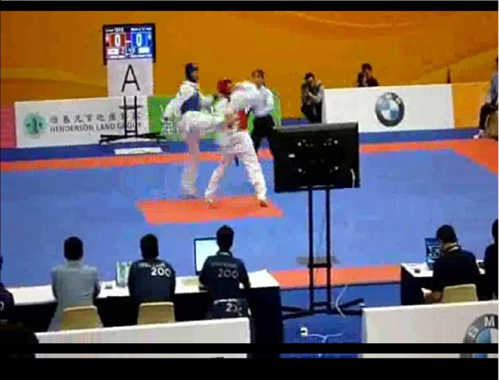 The image captures an intense moment at a martial arts tournament. Two athletes are in the midst of a combat forming a circle in the center of the ring, both wearing uniforms and protective gear. There is a referee visible, observing the match closely. The setting is an indoor sports arena with spectators seated around the court, which is marked blue and red, and a television monitor displaying '05-09' on the left side of the image, possibly indicating a time-keeping feature. The image also shows a watermark or logo at the bottom, likely representing the event organizer or sponsor.