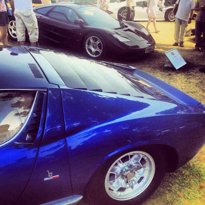 Post your Pictures here: Festival of speed 2013 - Page 1 - Goodwood Events - PistonHeads - The image showcases a vibrant outdoor scene featuring several vintage cars parked on dirt ground. Two cars, one black with a long, aerodynamic hood and the other blue, catch the eye. People are milling around the cars, suggesting this might be a gathering or event. The black car is slightly in the background, while the blue car is predominantly in the foreground, giving a sense of depth to the image.