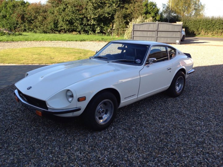 any 240z owners here? - Page 1 - Jap Chat - PistonHeads - The image depicts a pristine white Datsun 240Z sports car parked on a gravel driveway. The car's sleek design and chrome details are accentuated against the backdrop of a well-manicured lawn with verdant green grass and lush hedges. Adjacent to the driveway is a garage with a roll-up door closed, partially obscured by a tree. The overall scene exudes a sense of tranquility and serenity.