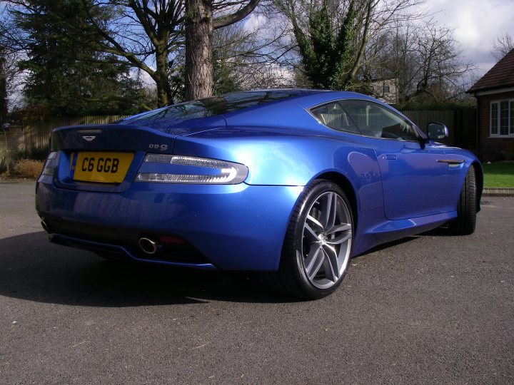 Can't wait till mid March!! - Page 2 - Aston Martin - PistonHeads - The image showcases a striking blue Aston Martin, which is parked on a blacktop surface. The car is a two-door coupe, featuring a shiny metallic paint job and a set of alloy wheels. Its license plate begins with "CS6". The backdrop includes a fence and the corner of a building, providing a contrast to the sleek beauty of the sports car.