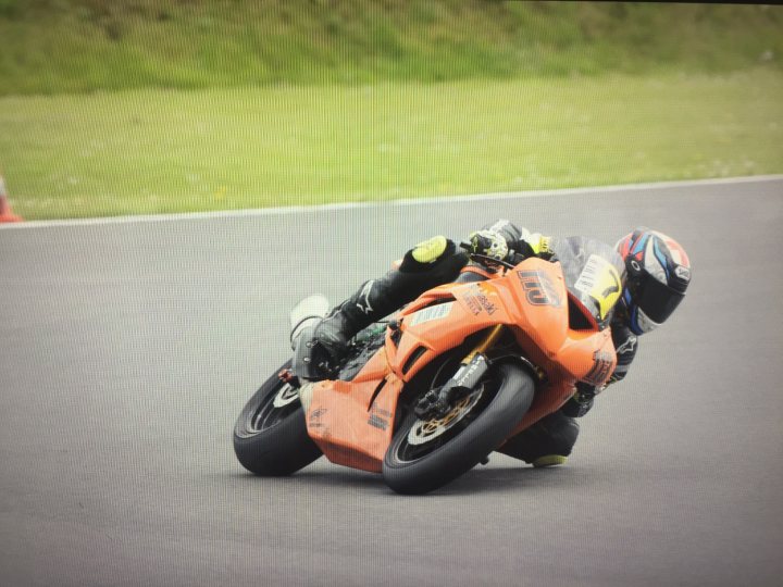 the bb trackday thread.   - Page 221 - Biker Banter - PistonHeads - The image captures a thrilling moment on a race track. A motorcyclist, dressed in a vibrant helmet and competitive gear, is leaning into a turn while riding an orange motorcycle. The rider's skillful body position and the bike's dynamic angle suggest high speed and precision control. The surrounding environment features a lush green landscape, providing a stark contrast to the adrenaline-fueled action of the race track. The perspective of the image appears to focus on the lower half of the motorcyclist, adding to the intensity and motion of the scene.