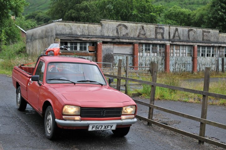 The Art of Shedding  - Page 31 - Readers' Cars - PistonHeads UK - In the image, a red pickup truck is traveling on a paved road. The truck is in motion, moving towards us from the background. Behind the truck, there's an old building that appears abandoned, with the sign "Garage" still visible. This suggests that this could have been a functional garage at one point, now left to decay. The landscape around the truck and the road is mostly flat with some greenery, indicating a rural or semi-rural area. There's no text other than the word "Garage" on the building.