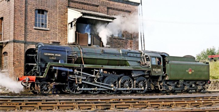 Pistonheads - The image shows a vintage steam locomotive pulling passenger cars at a train station. There are two trains visible, one in the foreground and another in the background, both with an old-fashioned design typical of steam engines. The locomotives appear to be in motion, with smoke billowing from their stacks. The setting includes railway tracks and infrastructure such as buildings and signage. The image conveys a nostalgic feeling, reminiscent of a bygone era of rail travel.