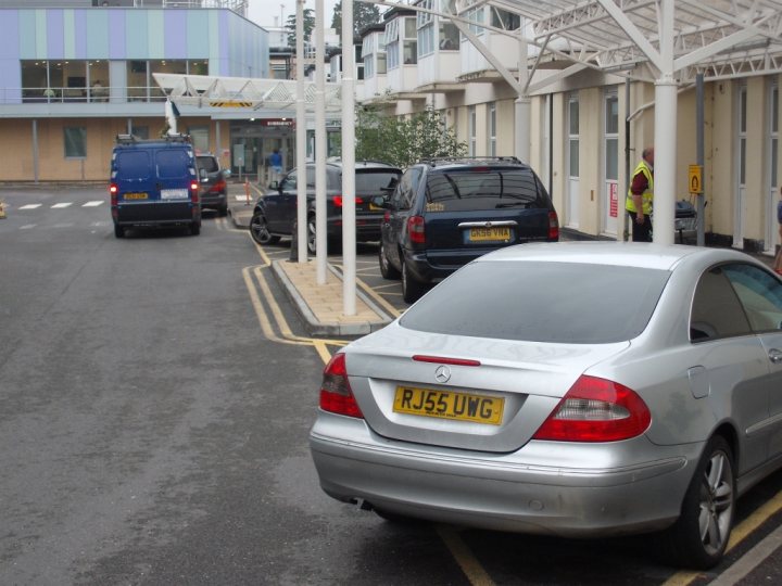 The BAD PARKING Thread [Vol 2] - Page 497 - General Gassing - PistonHeads - The image is a photograph taken during the day under overcast skies. It shows an outdoor scene featuring a row of parked cars on the side of a paved road. There are various makes and models, including a Mercedes-Benz, which has a yellow license plate with black letters and numbers. Another car next to it has a European-style license plate. People are visible in the background, with one individual near a building that appears to be a public transport stop or bus station. The design of the buildings suggests it could be a transit hub or an area with a concentration of businesses or services. The overall atmosphere is quiet and calm.