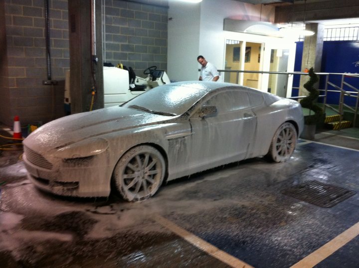 Pistonheads - The image depicts a car in a workshop that appears to have been involved in a painting or cleaning process. The car's hood and side surfaces are covered in fine particles, suggesting a stage of cleaning or paint removal. There is a man standing next to the car, likely supervising or inspecting the work. The setting seems to be inside a garage or workshop, with rails, pipes, and additional machinery in the background. The ground of the workshop has a patterned design and looks wet, which may be related to the work taking place on the car.